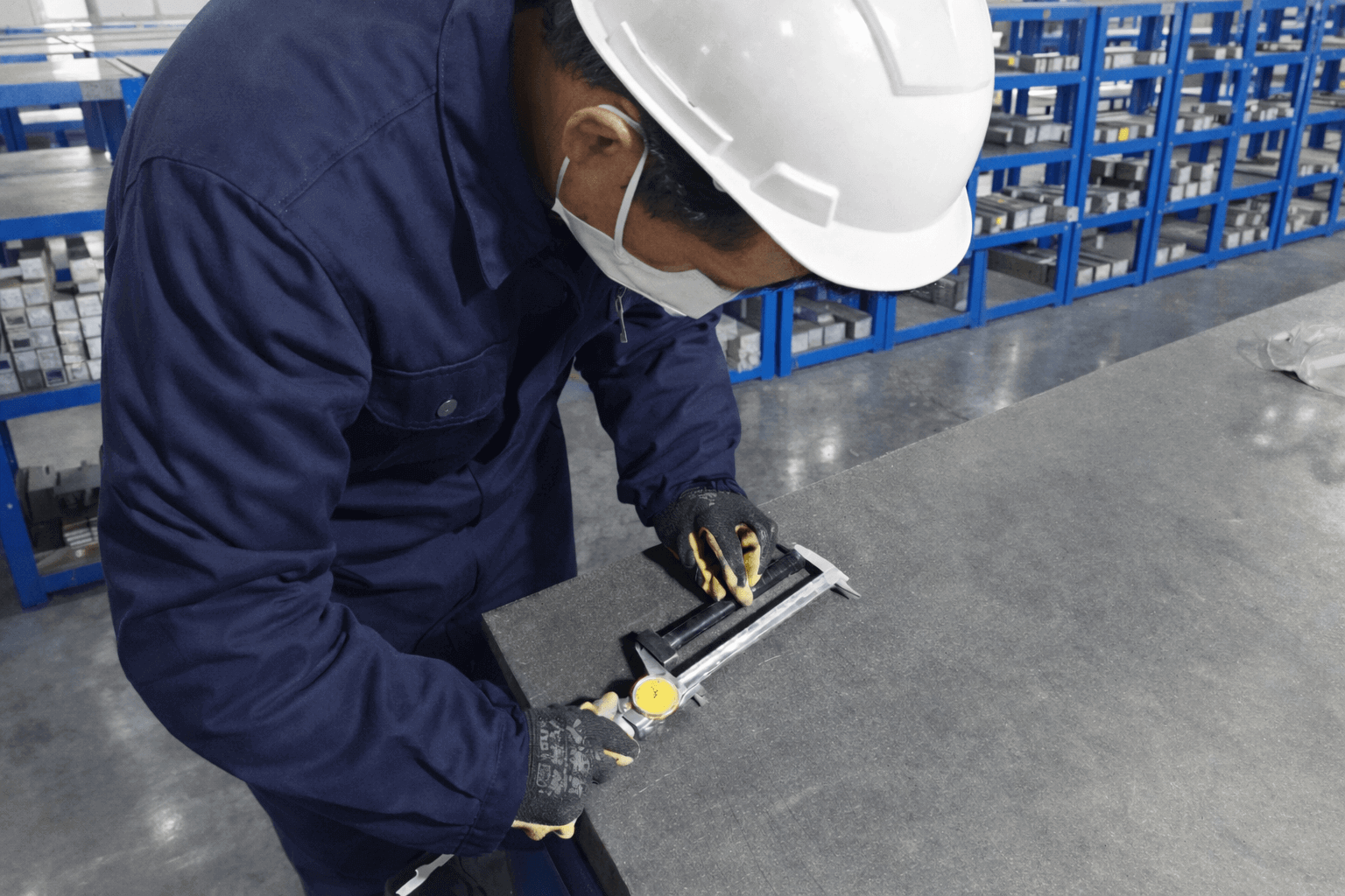 Factory worker wearing safety helmet and navy uniform measuring a metal component with a dial caliper on a granite inspection table