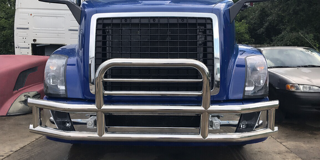 Heavy-duty stainless steel grille guard installed on a Class 8 semi truck providing front-end protection for headlights, radiator and bumper in fleet yard environment