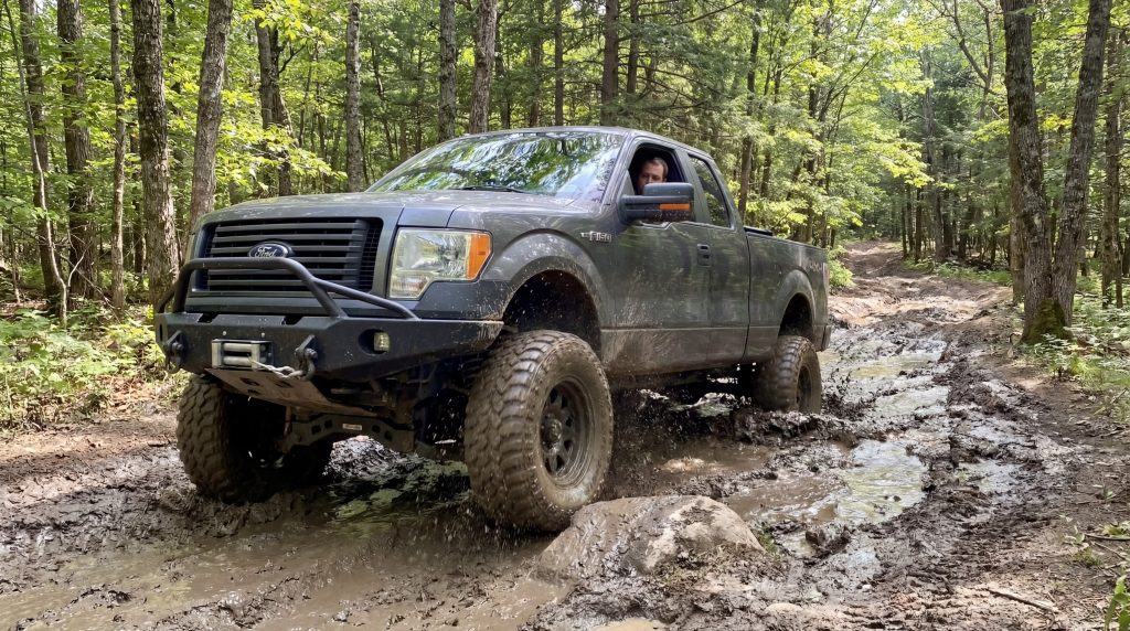 Off-road pickup truck driving through a muddy forest trail equipped with heavy-duty pickup bumpers designed for improved protection, recovery capability, and rugged terrain performance.