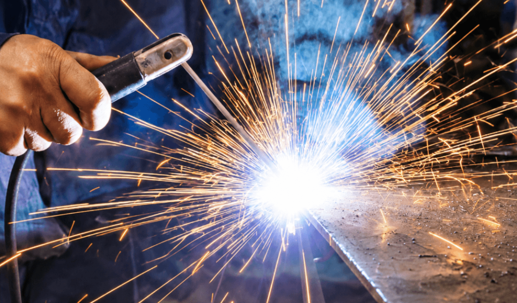 Close-up of a welder performing Stick Welding (SMAW) on a steel plate, with bright sparks and molten arc, in an industrial workshop environment.