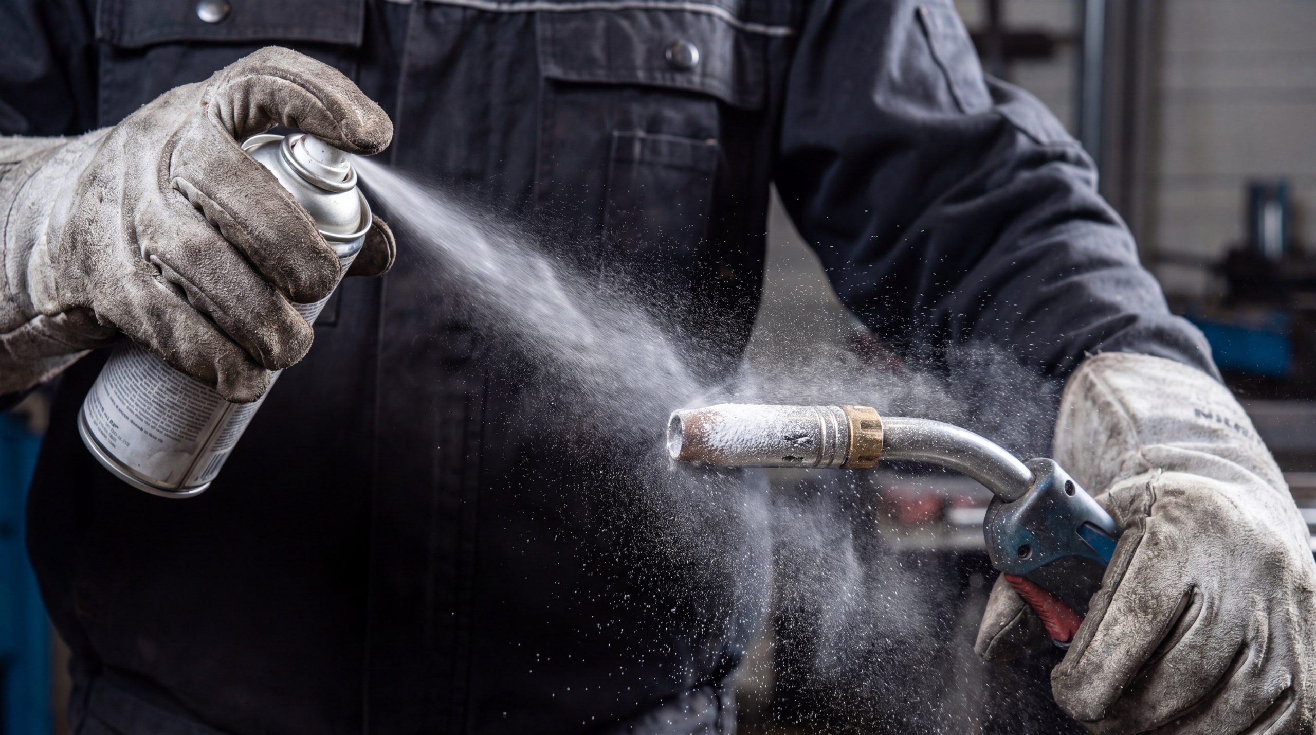 Professional welder applying a ceramic-based anti-spatter spray to a MIG welding nozzle to maintain gas flow.